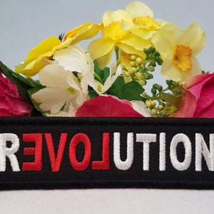 May include: Black embroidered patch with the word "REVOLUTION" in white and red lettering. The patch is surrounded by pink and yellow flowers.