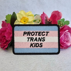 Może przedstawiać: Prostokątna naszywka z czarnym napisem "PROTECT TRANS KIDS" na jasnoniebieskim, białym i różowym tle w paski. Naszywka jest otoczona czarną obwódką i umieszczona na tle różowych, żółtych i zielonych kwiatów.