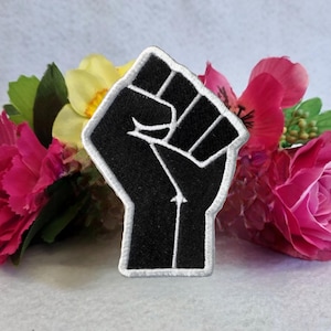 Puede incluir: Parche bordado en blanco y negro de un puño en alto con un contorno blanco. El parche está rodeado de flores rosas y amarillas.