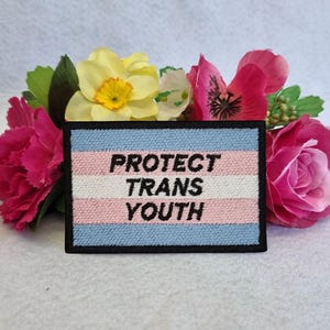 May include: A black and white embroidered patch with a transgender pride flag design. The patch says "Protect Trans Youth" in black text.