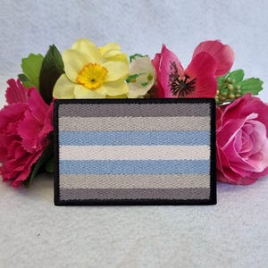 Könnte beinhalten: Ein grauer, weißer und hellblauer gestickter Patch mit schwarzem Rand. Der Patch ist rechteckig und hat fünf horizontale Streifen. Der Patch ist von rosa und gelben Blumen umgeben.