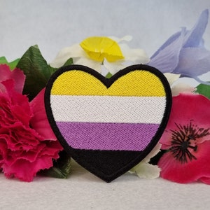 May include: A heart-shaped patch with the colors of the non-binary pride flag. The patch is yellow, white, purple, and black. It is surrounded by artificial flowers.