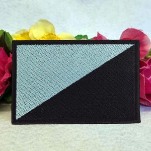 Könnte beinhalten: Ein gestickter Patch in Schwarz und Hellblau mit einer diagonalen Linie, die den Patch in zwei Hälften teilt. Die obere Hälfte ist hellblau und die untere Hälfte ist schwarz.