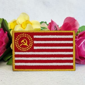 May include: Embroidered patch featuring a red and white striped design with a gold hammer and sickle emblem in the upper left corner. The patch has a gold border and is set against a backdrop of pink and yellow flowers.