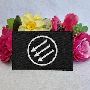 Może przedstawiać: Czarna łatka z tkaniny z białym kołem zawierającym trzy białe strzałki skierowane w prawo. Strzałki są ułożone po przekątnej.