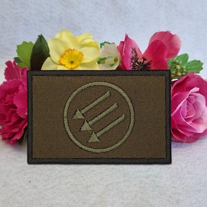 May include: A rectangular olive green embroidered patch featuring a dark green circular border enclosing three parallel diagonal arrows pointing downward to the left. The patch has a dark stitched edge and a textured fabric surface.