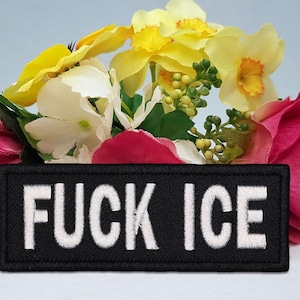 Pode incluir: Um patch retangular preto com as palavras "FUCK ICE" bordadas em branco, em um fundo de flores coloridas. As flores incluem rosas, amarelas e brancas.