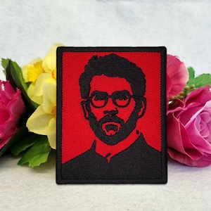 May include: A rectangular embroidered patch featuring a black portrait of a person with a beard and glasses set against a solid red background. The patch has a thick black border and is displayed in front of pink and yellow flowers.