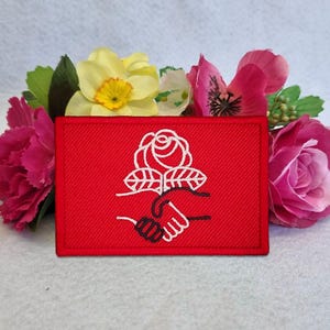 May include: A rectangular red embroidered patch featuring a white line art design of a rose positioned above two hands shaking. The patch has a stitched border and is displayed against a background of yellow, pink, and dark pink flowers.