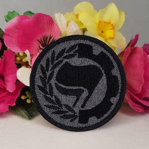 May include: Black and gray embroidered patch with a stylized fist in a circle surrounded by a wreath. The patch is on a background of pink and yellow flowers.