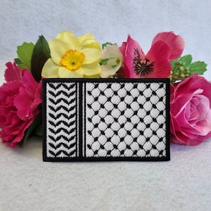 May include: Black and white embroidered patch featuring a traditional keffiyeh design. The patch is surrounded by pink and yellow flowers.