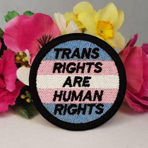 May include: A circular embroidered patch featuring the transgender pride flag colours of light blue, pink, and white horizontal stripes. The text "TRANS RIGHTS ARE HUMAN RIGHTS" is stitched in bold black lettering across the centre.