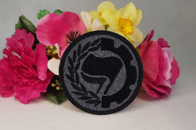 May include: Black and gray embroidered patch with a stylized fist in a circle surrounded by a wreath. The patch is on a background of pink and yellow flowers.