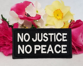 Puede incluir: Un parche rectangular negro bordado con el texto blanco NO JUSTICE NO PEACE en una fuente negrita sin remates. El parche tiene un borde cosido y está colocado frente a un grupo de flores artificiales de colores rosa, blanco y amarillo.