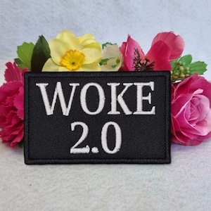 Op de afbeelding: Een zwart geborduurd embleem met de woorden "WOKE 2.0" in het wit. Het embleem is omgeven door kleurrijke bloemen, waaronder roze rozen, gele narcissen en andere bloesems.