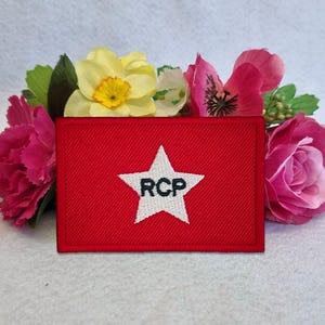 Puede incluir: Un parche rectangular rojo bordado que presenta una estrella blanca en el centro con las letras negras RCP cosidas en su interior. El parche está colocado frente a un grupo de flores artificiales amarillas y rosas.