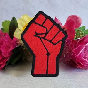 Könnte beinhalten: Ein roter, gestickter Aufnäher mit einer geballten Faust und schwarzem Umriss. Der Aufnäher ist vor rosa und gelben Blumen mit grünem Laub platziert, was einen auffälligen Farbkontrast erzeugt.
