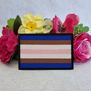 May include: A rectangular patch with a blue border and a striped design in blue, brown, pink, and white. The patch is set against a backdrop of pink and yellow flowers, creating a colourful contrast.