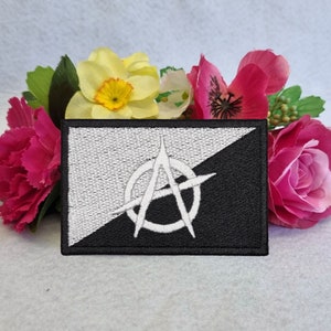 May include: Black and white embroidered patch with an anarchy symbol in the center. The patch is surrounded by a black border.