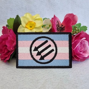 Może przedstawiać: Naszywka haftowana w kolorze czarnym i białym z trzema strzałkami w kółku na tle flagi dumy transpłciowej. Naszywka znajduje się na białym tle z różowymi i żółtymi kwiatami.