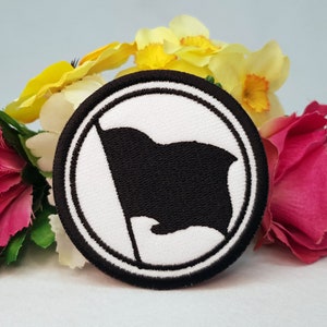 Könnte beinhalten: Ein weißer Kreis mit schwarzem Rand und einer schwarzen Flagge darin. Die Flagge weht an einem weißen Mast. Der Kreis ist von rosa und gelben Blumen umgeben.