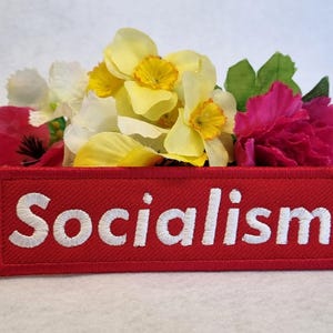 May include: A rectangular red embroidered patch featuring the word Socialism in white capital letters. The patch is positioned in front of a cluster of yellow, white, and pink flowers, showcasing a textured fabric finish and clear white stitching.