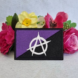 以下が含まれることがあります： 紫と黒の刺繍パッチに白い無政府主義のシンボルが描かれています。パッチは造花で囲まれています。