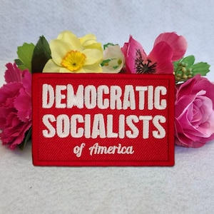 May include: A rectangular red embroidered patch featuring the white text "DEMOCRATIC SOCIALISTS of America" in a bold, sans-serif font for the first two words and a script font for the final two words, surrounded by pink and yellow flowers.