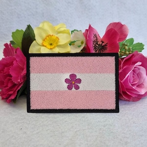 Puede incluir: Parche bordado en rosa, blanco y rosa claro con una flor morada en el centro. El parche tiene un borde negro.