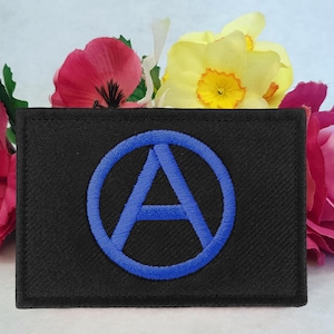Pode incluir: Um emblema retangular bordado em preto com um símbolo circular azul centralizado contendo uma letra A maiúscula. O emblema possui uma borda costurada e uma superfície plana, exibindo um design limpo com fios de alto contraste em azul e preto.