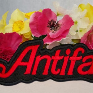 Pode incluir: Um emblema bordado preto com a palavra Antifa em uma fonte cursiva vermelha em negrito com uma borda preta. O emblema está posicionado na frente de um grupo de flores artificiais em tons de rosa, amarelo e branco, incluindo rosas e papoulas.