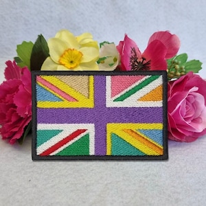 May include: Embroidered patch featuring a colorful, stylized Union Jack design. The patch has a black border and incorporates various colors, including purple, yellow, pink, green, and blue. The patch is surrounded by pink and yellow flowers.