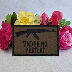 Puede incluir: Un parche rectangular marrón con una silueta de rifle negro y el texto "UNDER NO PRETETX" en negro. El parche está rodeado de flores rosas y amarillas con hojas verdes.