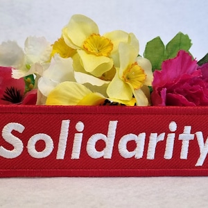 Peut inclure: Un écusson rectangulaire rouge brodé avec le mot Solidarity en lettres capitales blanches. L'écusson est placé devant un bouquet de fleurs artificielles, comprenant des jonquilles jaunes et des œillets roses, avec une bordure cousue.