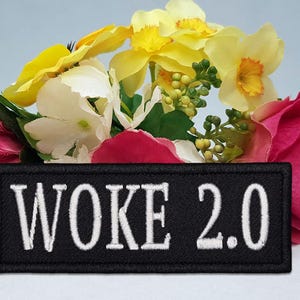 May include: A black rectangular patch with the text "WOKE 2.0" embroidered in white, set against a backdrop of colorful flowers. The flowers include yellow, white, and pink blooms.