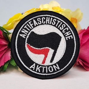 May include: A circular embroidered patch featuring a black and red flag design on a white background, enclosed by a thick black border. The text ANTIFASCHISTISCHE AKTION is stitched in white capital letters along the top and bottom edges.