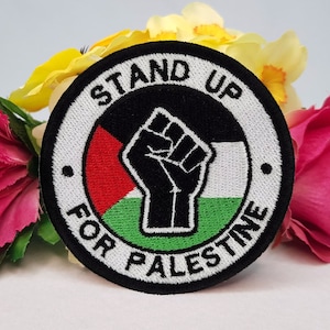 May include: A black and white embroidered patch with a raised fist in the center. The fist is surrounded by a red, white, and green Palestinian flag. The text "Stand Up For Palestine" is written in a circle around the fist.