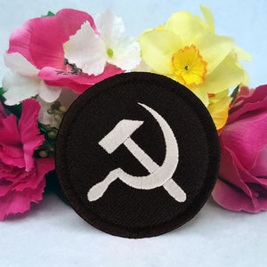 May include: A white embroidered hammer and sickle symbol on a black circular patch. The patch is surrounded by pink, white, and yellow artificial flowers.