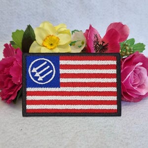 Può includere: Una toppa rettangolare ricamata con un design ispirato alla bandiera americana con strisce orizzontali rosse e bianche. Il cantone blu contiene un cerchio bianco che racchiude tre frecce diagonali rivolte verso l'angolo in basso a destra.