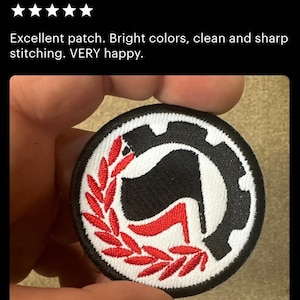 May include: A black and white embroidered patch with a red stylized 'A' inside a circle. The 'A' is surrounded by a red wreath. The text 'Antifa (anti-fascist) wreath' is visible above the patch.