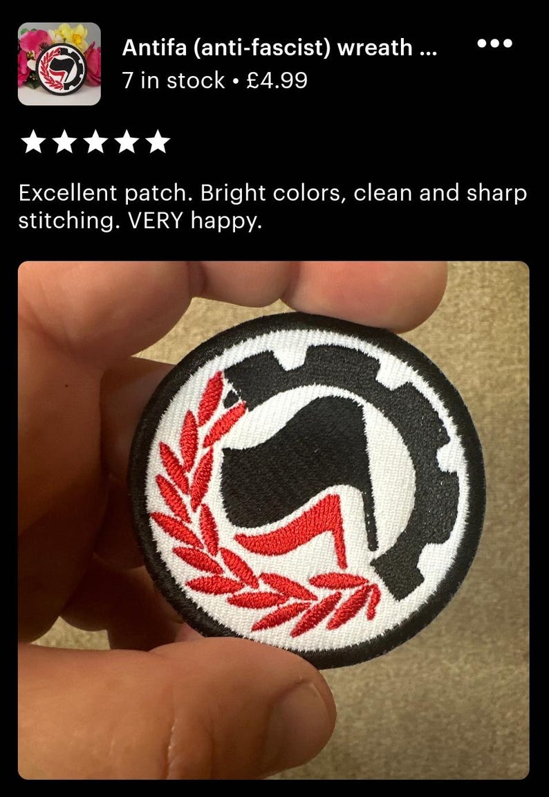 May include: A black and white embroidered patch with a red stylized 'A' inside a circle. The 'A' is surrounded by a red wreath. The text 'Antifa (anti-fascist) wreath' is visible above the patch.
