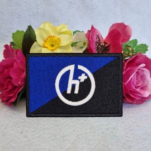 Può includere: Una toppa ricamata blu e nera con un cerchio bianco che contiene la lettera "H" e un segno più. La toppa è circondata da un bordo nero.