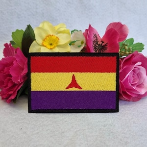 May include: An embroidered patch featuring a red, yellow, and purple flag with a red triangle in the center. The patch is surrounded by a black border.
