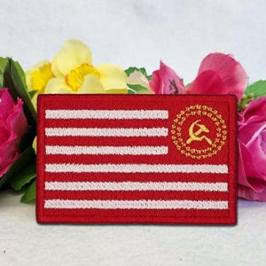 May include: An embroidered patch in red and white, designed to look like a flag. It has white stripes and a gold emblem of a hammer and sickle inside a circle. The patch is set against a backdrop of pink and yellow flowers.
