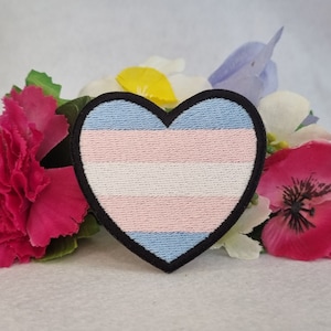May include: A heart-shaped patch with the transgender pride flag colors: light blue, pink, white, pink, light blue. The patch is surrounded by artificial flowers.