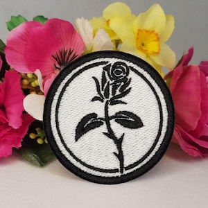 Op de afbeelding: Geborduurde patch met een zwart rozenontwerp in een witte cirkel, omlijnd door een zwarte rand. De patch is geplaatst tegen een achtergrond van helder roze, gele en witte bloemen, wat een opvallend contrast creëert.