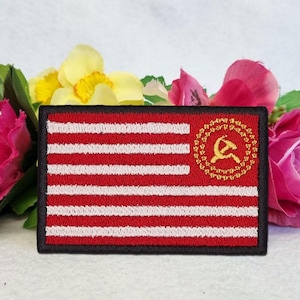 May include: Embroidered patch with a red and white striped design and a gold hammer and sickle emblem. The patch has a black border. The image is set against a backdrop of pink and yellow flowers.