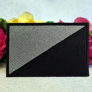 Könnte beinhalten: Ein gestickter schwarzer und grauer Patch mit einer diagonalen Linie, die den Patch in zwei Hälften teilt. Die obere Hälfte ist grau und die untere Hälfte ist schwarz.