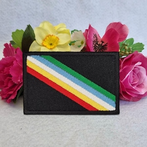 Op de afbeelding: Een zwart rechthoekig embleem met een diagonaal streepontwerp in rood, geel, wit, blauw en groen. Het embleem wordt omringd door roze en gele bloemen.