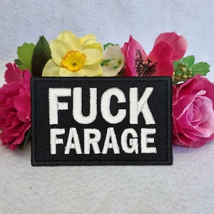 以下が含まれることがあります： 「FUCK FARAGE」の白い文字が刺繍された黒い長方形のパッチ。パッチは、ピンク、黄色、緑色の造花のアレンジメントで囲まれています。